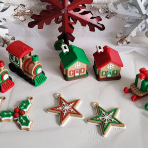 Tabletop arrangement of small Christmas ornaments and cookies on white cloth against a white brick wall, featuring two red-and-green toy trains, two miniature houses with green and red roofs, two sleds carrying wrapped presents, holly and star-shaped decorated cookies, large red and white snowflake decorations and pinecones.