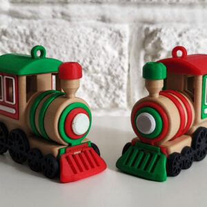 Two small beige toy steam locomotives with black wheels and red-and-green accents (one with a green roof and red cowcatcher, the other red-roofed with a green cowcatcher) sit side by side on a white surface in front of a textured white brick wall.
