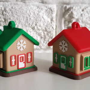 Two small plastic holiday house ornaments side by side on a white surface in front of a textured white brick wall — one with a green roof and snowy chimney and red-trimmed windows, the other with a red roof and green-trimmed windows, both bearing a snowflake decoration and a hanging loop on top.