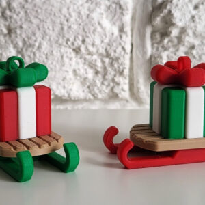 Two small wooden sled ornaments on a white tabletop against a textured white brick wall, each carrying a striped red, white and green gift topped with a matching bow (left sled has green runners, right sled has red runners).