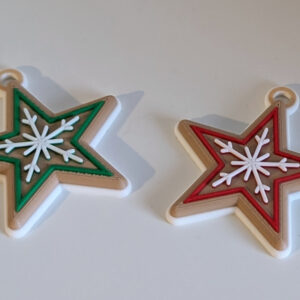 Two star-shaped Christmas ornaments with beige edges and white snowflake patterns—one with green inlay and one with red inlay—lying on a white surface beneath a textured white brick wall.