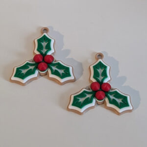 Two small holly-shaped Christmas ornaments with three green leaves outlined in white and three red berries, each with a top hanging loop, placed on a white surface against a textured white brick background.