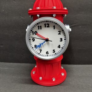 Decorative red fire-hydrant-shaped clock with a round white face, black numerals and silver bezel and bolts, featuring a red hand, a black hand and a blue splash-shaped hand, set against a plain dark gray background.