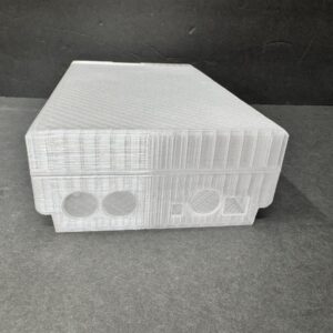 Translucent 3D-printed rectangular box with visible layer lines and textured top, showing two circular cutouts and a larger circular plus a small square cutout on the front edge, sitting on a dark gray surface against a black background.