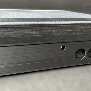 Close-up of a black rectangular electronic enclosure angled to show its front edge with three circular multi-pin DIN connectors, a small blue component visible through a cutout, and textured horizontal grooves across the metal surface.