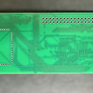 Green rectangular printed circuit board lying face-down on a blurred gray surface, showing etched trace routing, a square array of plated through-holes on the left, a long row of plated holes along the top, and a semicircular notch plus a round mounting hole at the right edge.