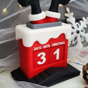 Small red decorative countdown calendar shaped like a snow-topped chimney with two black-and-red Santa legs sticking out the top and the numbers 31 displayed beneath the words “Days Until Christmas,” set on a black base and surrounded by pinecones, a white wooden snowflake, tulle and fairy lights.