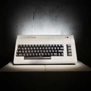 Beige Commodore 64 home computer with rainbow logo, front-facing on a lit pedestal against a dark textured wall, showing its black keycaps and column of function keys.