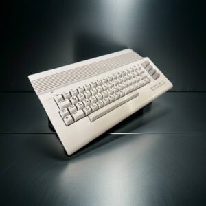 Retro beige home computer keyboard with chunky keycaps, horizontal ridges on the top panel and four vertical function keys on the right, photographed at an angle on a dark reflective metal surface.