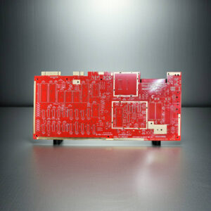 Red unpopulated printed circuit board with white silkscreen markings and gold contact pads standing upright on a reflective metallic surface against a soft gray gradient background.