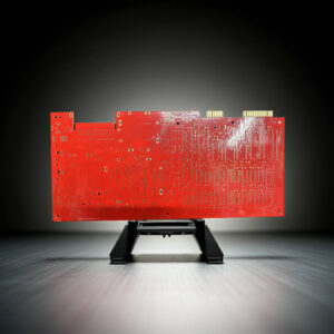 Red printed circuit board with gold edge connectors and numerous through-hole pads and traces mounted vertically on a small black stand against a gradient gray background