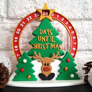 Christmas countdown decoration featuring a green tree with gold "DAYS UNTIL CHRISTMAS" text and a red numbered circular dial topped by a gold star showing 24, a cartoon reindeer and two small decorated trees on a white base, flanked by pine cones and white ornaments against a white brick background.