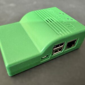 Small matte green plastic electronics enclosure resting on a dark surface, showing two stacked USB-A ports, an Ethernet jack and a micro‑USB power port on one side, with ridged vents and embossed lettering on the top.