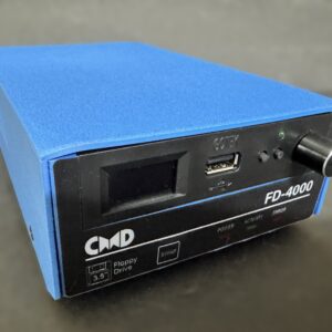 Blue box-shaped external device with a black front panel labeled FD-4000 and CMD, showing a small rectangular display, a USB port, a rotary knob, status LEDs and markings for a 3.5" floppy drive, resting on a dark surface.