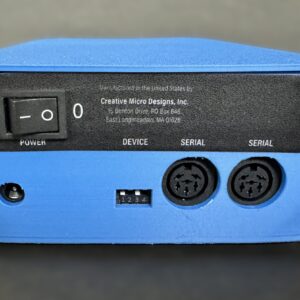 Front view of a small blue electronic box featuring a black rocker power switch labeled 1/0, a DC power jack, a four-position DIP switch, and two round five-pin serial (DIN) connectors under a black label reading Creative Micro Designs, Inc.