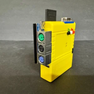 Small yellow electronic module standing upright on a dark surface, showing three stacked round DIN connectors (green, black, purple) on its side, colored indicator LEDs and a metal serial-style port on top, with a plain gray background.